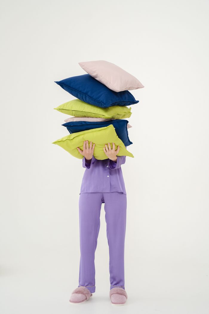 Person in purple pajamas holding a stack of colorful pillows in a studio setting.